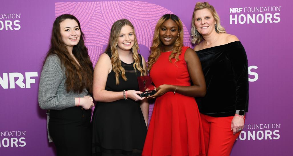 Four individuals stand together in front of a purple backdrop featuring the text 'NRF Foundation Honors.' They are dressed in formal attire, with one person holding an award. The backdrop prominently displays the NRF Foundation logo and the word 'HONORS' repeated multiple times, emphasizing the recognition of achievements at this formal event.
