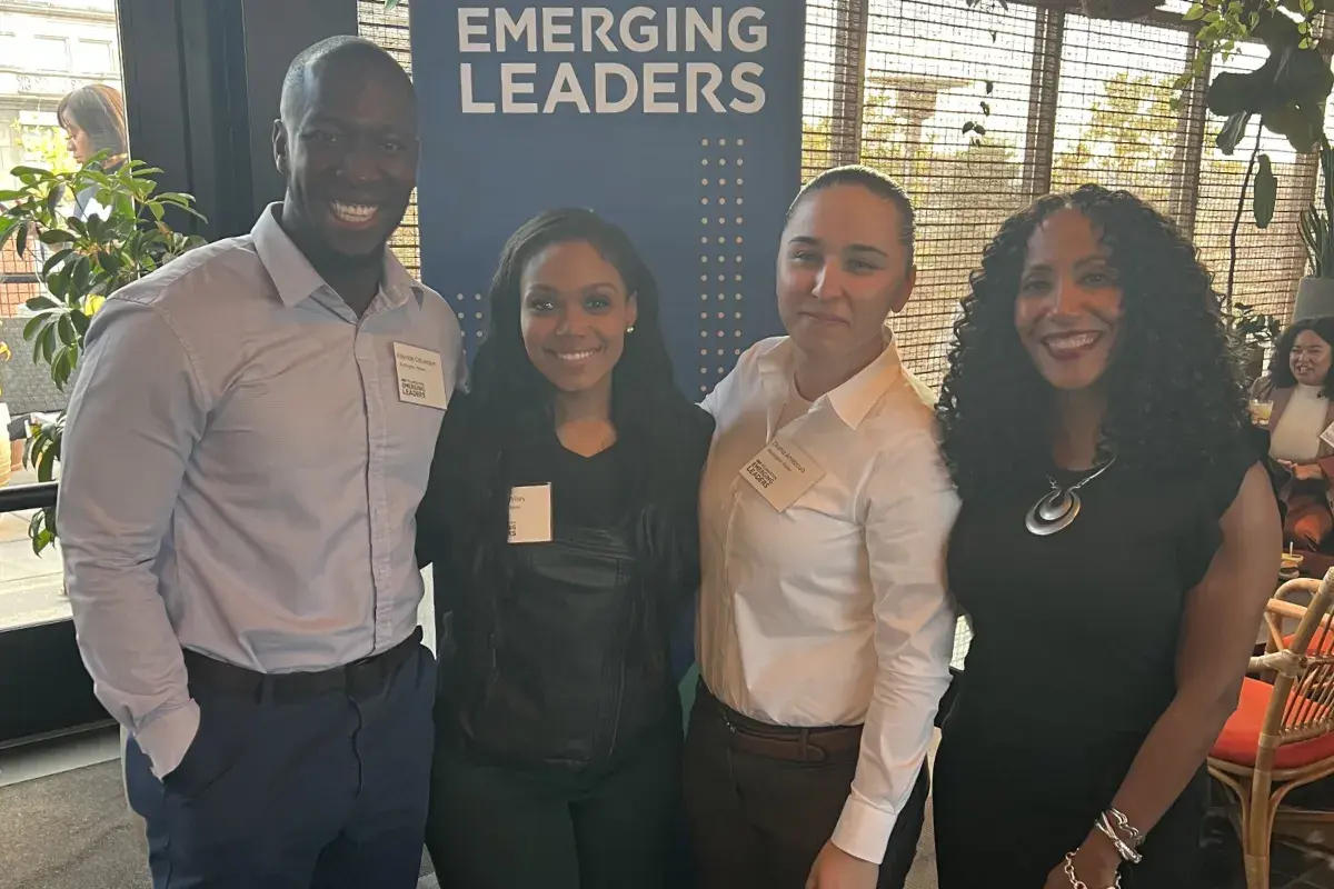 A group of four people standing together at an event beside a vertical banner that reads NRF Foundation Emerging Leaders. They are indoors in a space with plants, natural light, and seating visible in the background.